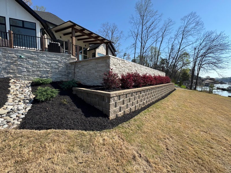 A stone retaining wall with a seat wall, red shrubs in fresh mulch, and a lake view beside a home with a covered porch