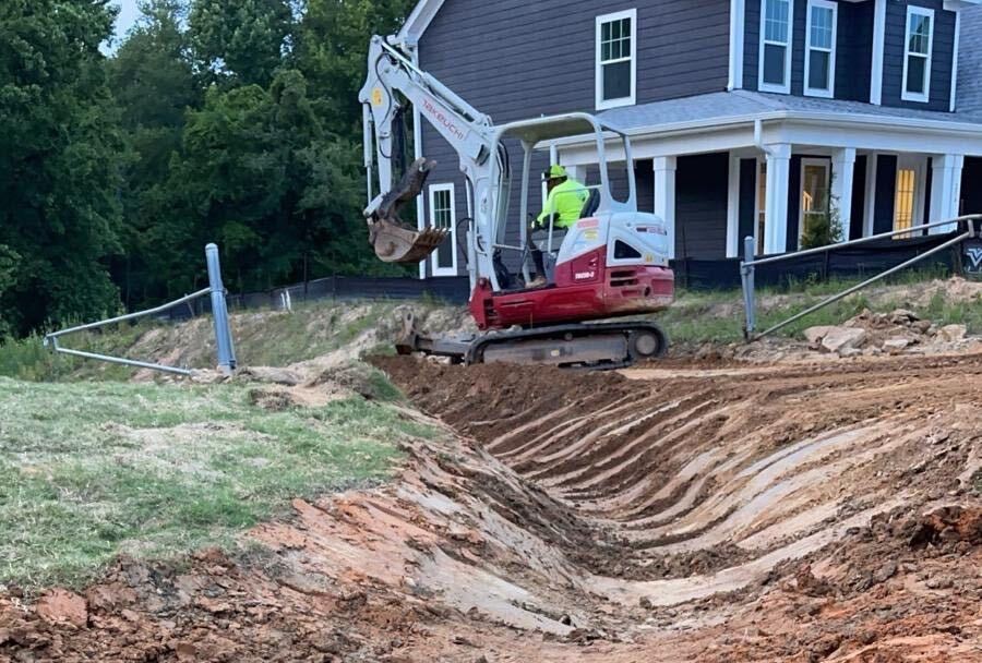 A compact excavator cutting a curved drainage swale beside a home, with graded banks and sculpted soil leading toward the machine.